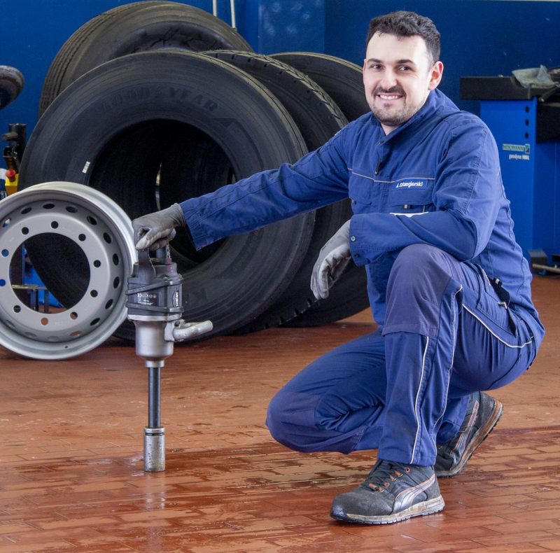 Lkw Reifen kaufen bei BTS. Lkw Monteur mit Werkzeug in hockender Position vor aufgereihten Lkw Reifen und Reifen für Auflieger sowie einer Lkw Felge