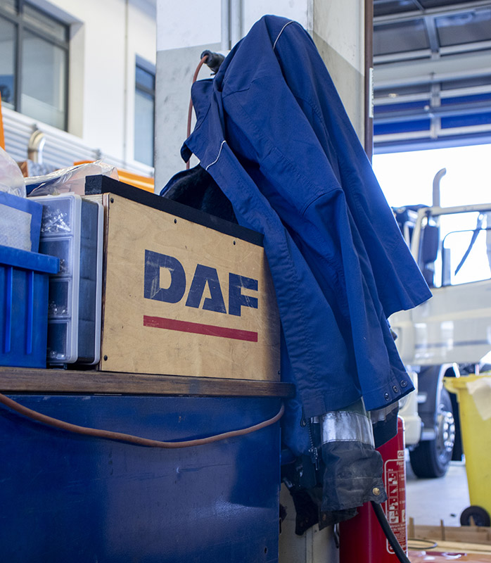 BTS Lkw-Werkstatt. Eine dunkelblaue Arbeitsjacke hängt über einer Kiste mit dem Logo von DAF.